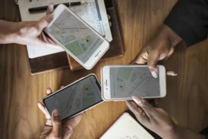 An overhead view shows three people holding smartphones over a wooden table with open notebooks. Each screen displays a navigation app with a map and a red location pin, suggesting they are coordinating a route or destination together.