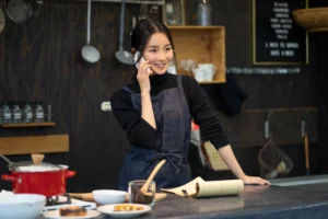 A smiling woman wearing a dark turtleneck and a denim apron stands behind a kitchen counter while talking on a white smartphone. In front of her, a notepad with a pen rests on the counter alongside small bowls of food and a red pot on a stove.