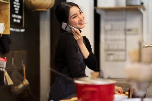A smiling Asian woman wearing a dark long-sleeved shirt and a denim apron stands in a restaurant or kitchen while talking on a white smartphone. Her other hand rests on the counter near a red pot, with shelves and kitchen items softly blurred in the background.