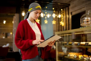 A person wearing a grey headscarf and a bright red cardigan stands in a bakery or café while looking down at a menu. In the foreground, a glass display case is filled with various pastries and cakes with small price tags.