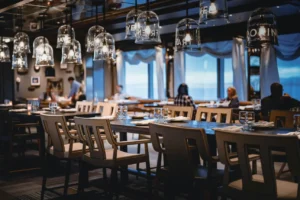 The image shows the interior of a dimly lit, upscale restaurant featuring long wooden tables set with glassware and modern beige chairs. Unique glass bell jar pendant lights with white text hang from the ceiling, while patrons sit at tables in the blurred background near large windows overlooking the sea.