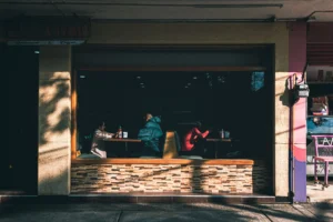 A view from the street looks through a large window into a dimly lit restaurant where patrons are seated at booths, framed by a textured stone base and warm overhead lighting. The composition closely mirrors Edward Hopper’s famous painting, Nighthawks, capturing a moody, cinematic slice of urban life.