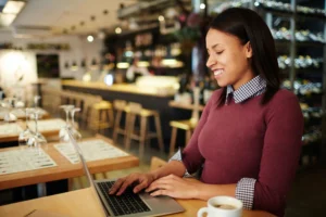 In the image, a smiling woman sits at a wooden table in a cafe or restaurant while typing on a laptop. She is wearing a maroon sweater over a collared gingham shirt, with a coffee cup resting nearby against a softly blurred background of the venue's interior.