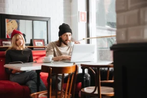 A man in a black beanie sits at a cafe table, typing on a laptop and holding a white coffee cup. Beside him on a red booth, a woman wearing a red beanie sits reading a book or magazine.