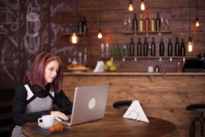 A woman with reddish hair and glasses sits at a wooden table in a cozy coffee shop, focused on working on her laptop. In front of her sits a white coffee cup and a croissant on a small slate plate, with a rustic bar and shelves of bottles blurred in the background.