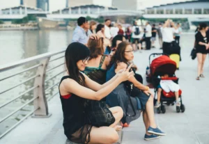 A woman with long black hair and glasses sits cross-legged on a waterfront ledge, holding up her phone to take a photo or selfie. In the background, a crowd of people, including someone with a red stroller, relaxes along the bustling boardwalk.