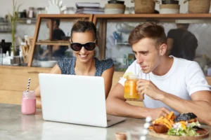 In the image, a smiling woman wearing sunglasses and a man sipping a jar of juice sit together at a cafe counter while looking at an open laptop. They are surrounded by snacks, including a pink smoothie, a burger, and fries, with a bakery display counter visible in the background.