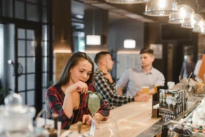 In the image, a young woman wearing a red plaid shirt sits at a wooden bar counter and looks down at a green cocktail garnished with mint. In the softly blurred background, two men are seated further down the bar, chatting over beers under hanging glass lights.