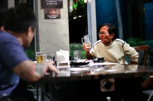 An older man with glasses raises a mostly empty beer mug while sitting at a restaurant table, appearing to talk mid-sentence. In the foreground, another man with a tattooed arm is seen from the back, listening across the table.