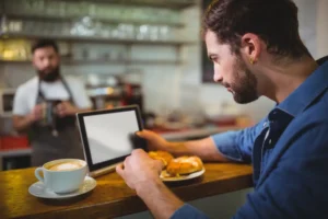 A man with a beard and ear gauge rests at a wooden cafe counter, looking intently at a blank tablet screen while holding a pastry. In the blurred background, a barista in an apron stands behind the bar near a cup of latte art coffee.