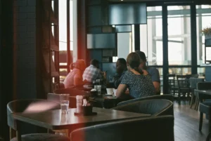 A group of people are seated at tables inside a restaurant or cafe with large windows looking outside. The image has a grainy, film-like quality with a prominent red light leak stretching across the left side of the frame.