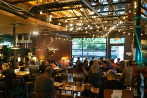 The image shows the bustling interior of a lively brewpub or restaurant filled with patrons sitting at wooden tables and a long bar. The space features industrial decor with an exposed wooden ceiling framework, numerous hanging string lights, and a large glass garage-door window looking out onto a street.