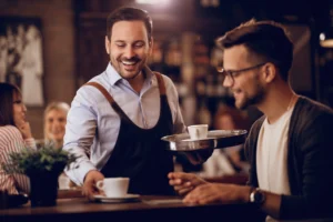 In the image, a smiling male waiter wearing an apron serves a cup of coffee to a seated male customer at a cafe counter. The warm, dimly lit background features other patrons enjoying the cozy atmosphere of the establishment.