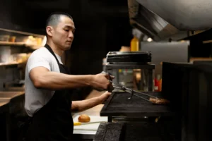 An Asian male chef wearing a white t-shirt and black apron uses long metal tongs to flip a burger patty on a commercial grill. The dim kitchen environment features stainless steel countertops, a cutting board with a bun, and a large ventilation hood overhead.