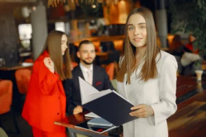A smiling young woman in a white blazer stands in the foreground of a cafe, holding an open black binder and looking toward the camera. In the blurred background, a man in a suit and another woman in a bright red blazer sit at a table, looking on.