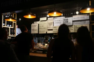 The image shows a group of people viewed from behind standing at a dimly lit Portuguese food counter. They are looking up at a large menu board illuminated by warm, overhead pendant lights displaying various dishes like tapas, fish, and burgers.