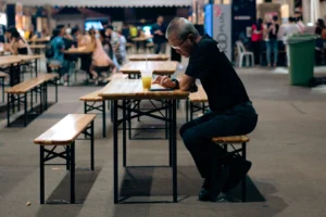 An older man in a black shirt and pants sits on a wooden bench at an outdoor food court, leaning over a table to use his smartphone. In front of him is a plastic cup with a yellow drink and a straw, while the blurred background shows other people dining at similar wooden tables.