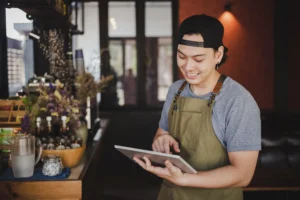 The image shows a smiling young Asian barista standing inside a coffee shop. He wears a green apron over a grey t-shirt and a backward black baseball cap while interacting with a digital tablet in his hands.