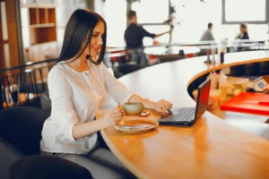 A young woman with long, straight dark hair smiles while sitting at a curved wooden bar counter and working on a laptop. She holds a coffee cup over its saucer with one hand and rests her other hand near the laptop's keyboard.