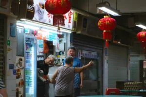 An older woman with her back to the camera gestures toward two men working at a traditional hawker center food stall. Red Chinese lanterns hang from the ceiling above the illuminated beverage refrigerator and menu boards.