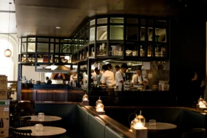An open-concept restaurant kitchen is bustling with several chefs in white uniforms preparing food behind a dark, sleek counter. The foreground features an upscale, dimly lit dining area with empty tables, green leather booths, and warm, glowing tabletop lights.