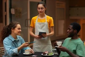 A server wearing a yellow shirt and apron stands at a cafe table taking an order on a tablet from two seated customers. A woman on the left holds a smartphone near her coffee cup, while a man on the right smiles and holds a teacup near a plate of cookies.