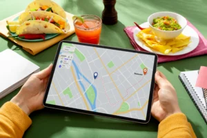 A pair of hands holds a tablet displaying a map route to a restaurant on a green table. Surrounding the tablet is a spread of Mexican food, including tacos, guacamole with tortilla chips, and a refreshing cocktail.