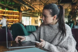 A woman with her hair tied back in a ponytail sits at a wooden table in a dimly lit café, typing on an open laptop. She wears a cozy grey knit sweater and holds a white smartphone in her other hand while focusing attentively on the screen.