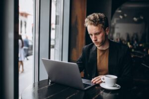 A young man with curly blonde hair sits at a café table next to a large window while working focused on his laptop. A white coffee cup on a saucer rests on the dark wood table beside him as people walk by on the street outside.