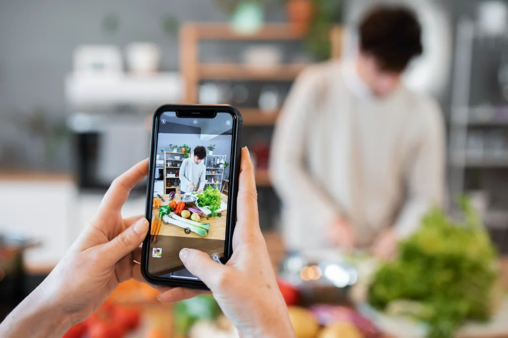 A person holds up a smartphone to record a man preparing fresh vegetables at a wooden kitchen counter. The phone screen clearly captures the man chopping green herbs alongside a colorful spread of peppers, carrots, and leeks.