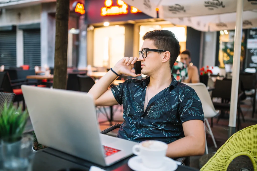 A young man with glasses and a patterned short-sleeved shirt sits at an outdoor cafe table talking on his smartphone. In front of him on the table are an open laptop and a cup of coffee on a saucer.