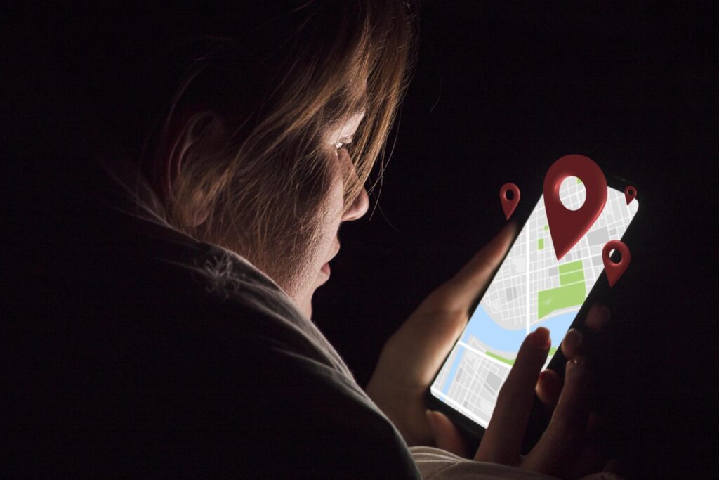 A woman looks at a glowing smartphone screen in a dark room while holding the device with both hands. The screen displays a digital map interface with prominent red location pin icons floating above it.