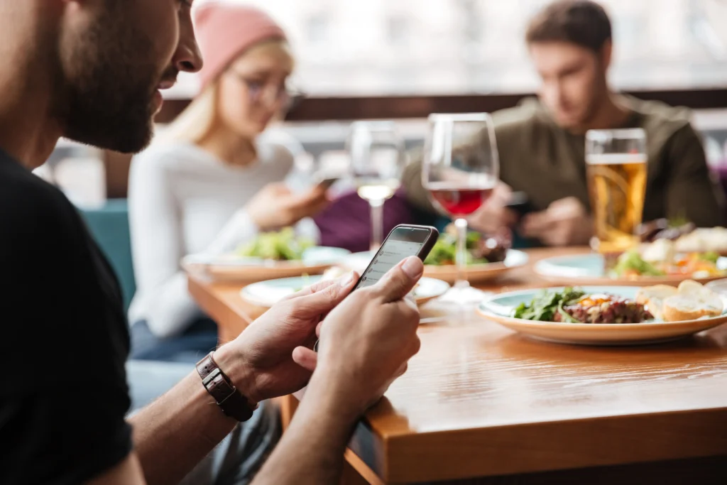 A side view of a man holding and looking at his smartphone while seated at a dining table. In the blurred background, two friends are also absorbed in their phones over a meal with drinks.