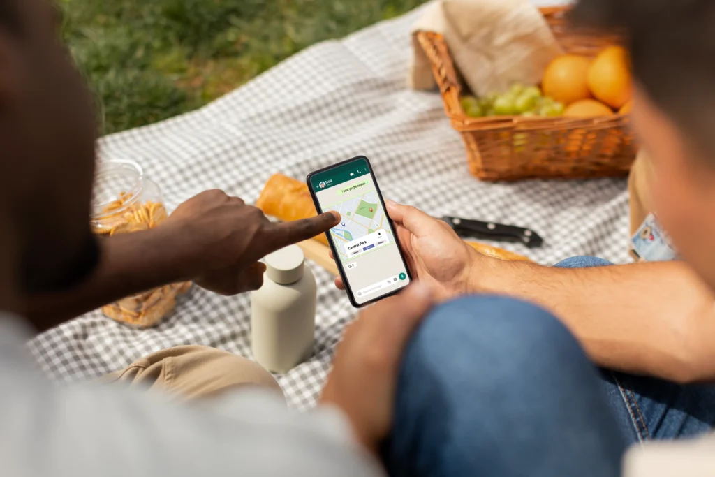 Two people sit outdoors on a checkered picnic blanket while looking at a navigation map on a smartphone. One person holds the phone while the other points directly at a location pin on the screen.