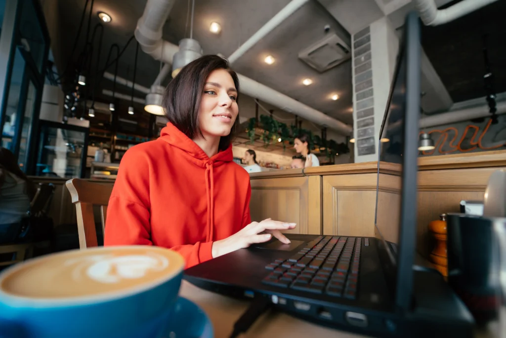 A young woman with short dark hair wearing a bright red hoodie works on a laptop at a cafe table. In the foreground, a blue cup of coffee with latte art sits on the wooden table beside her.