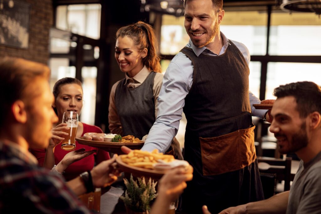 Two smiling servers deliver platters of french fries and sausages to a group of friends seated at a restaurant table. The guests enjoy drinks and good conversation in a cozy, warmly lit dining atmosphere.