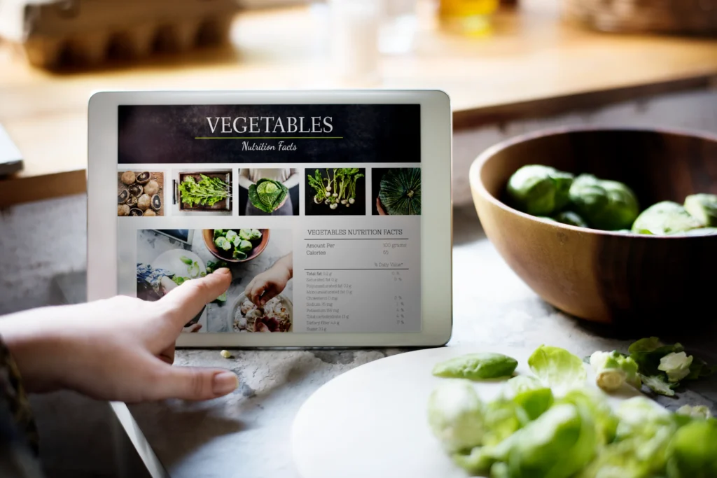A hand points to a digital tablet displaying vegetable nutrition facts on a kitchen counter. Beside the device, fresh Brussels sprouts rest in a wooden bowl and on a cutting board nearby.