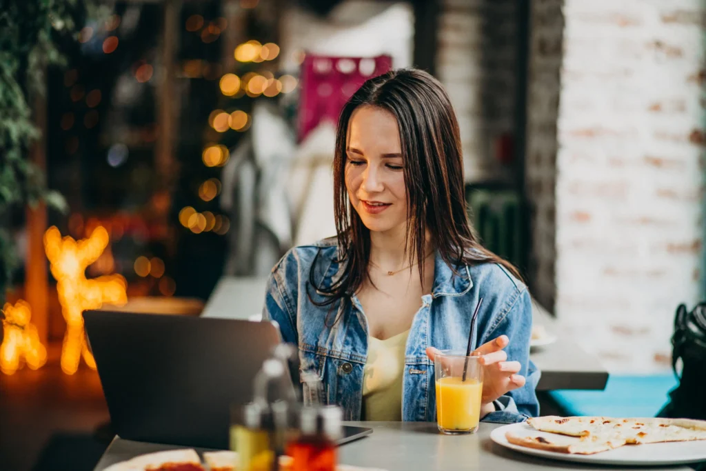 A young woman in a denim jacket smiles as she looks at her open laptop while sitting at a table in a cozy restaurant or café. On the table before her are a glass of orange juice with a straw and plates of flatbread or pizza, with warm bokeh lights glowing in the background.