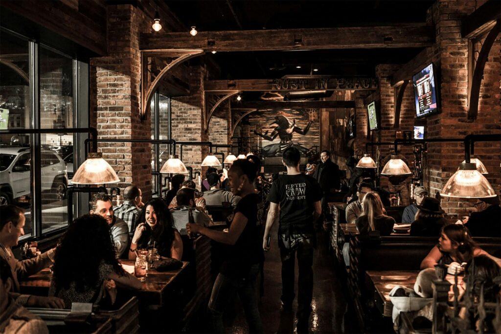 A bustling, dimly lit restaurant features rustic brick walls, exposed wooden beams, and warm pendant lighting above occupied booths. Patrons chat and dine while staff wearing black shirts move down the central aisle toward a large mural in the background.