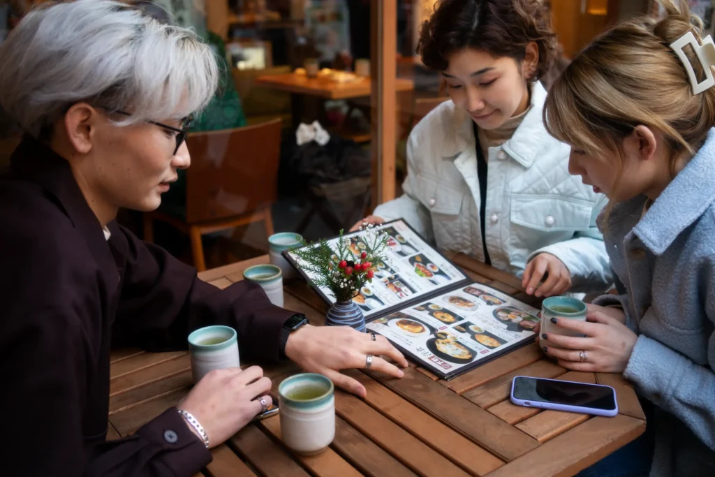 Three young friends sit together at a wooden outdoor table, looking over an open menu to choose their meal. Small ceramic tea cups and a tiny floral arrangement rest on the table between them.