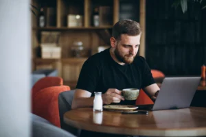 A bearded man in a black t-shirt sits at a wooden cafe table while focused on his laptop screen. He holds a coffee cup in his hand next to a small bottle of milk and a plate of food.