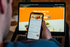 A person holds a smartphone displaying a food delivery app with a McDonald's menu open on the screen. In the background, a open laptop displays a matching food ordering website with an orange header.