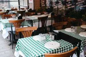 An outdoor dining area features wooden chairs and tables set with green-and-white checkered tablecloths. Each table is neatly arranged with white plates, inverted drinking glasses, and napkin dispensers.