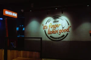 A dimly lit KFC fast-food restaurant interior featuring a wall adorned with the illuminated slogan, "it's finger lickin' good." To the left, an orange "ORDER HERE" sign hangs above a dark ordering kiosk counter.
