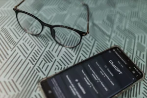 A smartphone displays the ChatGPT homepage interface in dark mode next to a pair of black-framed eyeglasses. Both items rest on a light green patterned surface with a repeating geometric line design.