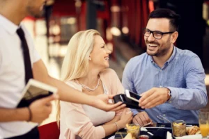 A smiling man pays for lunch at an outdoor restaurant by tapping his smartphone on a payment terminal held by a waiter. His female partner looks on happily as they finish their meal.
