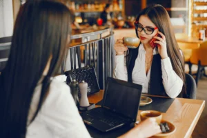 Two stylish women sit at a cafe table equipped with an open laptop, coffee cups, and a black handbag. One woman wearing glasses holds a cup to her mouth while talking on her phone as her companion sits across from her.