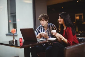 A man in a plaid shirt and a woman in a red top sit together at a dark table in a cozy café or restaurant, both focused on an open laptop. They hold small white coffee cups as they intently view the screen, with a flower vase and candle decorating the table beside them.