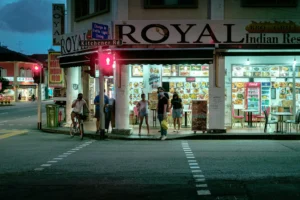 A street corner scene at dusk shows pedestrians and a cyclist wearing face masks waiting at a crosswalk outside the brightly lit "ROYAL Indian Restaurant." A red pedestrian light illuminates the corner at the intersection of Kitchener Road, where menu displays fill the restaurant windows.