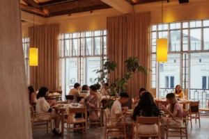 People dine at round wooden tables inside a warm, brightly lit cafe with high ceilings. Large multi-pane windows lined with beige drapes and glowing yellow pendant lamps create a cozy atmosphere.